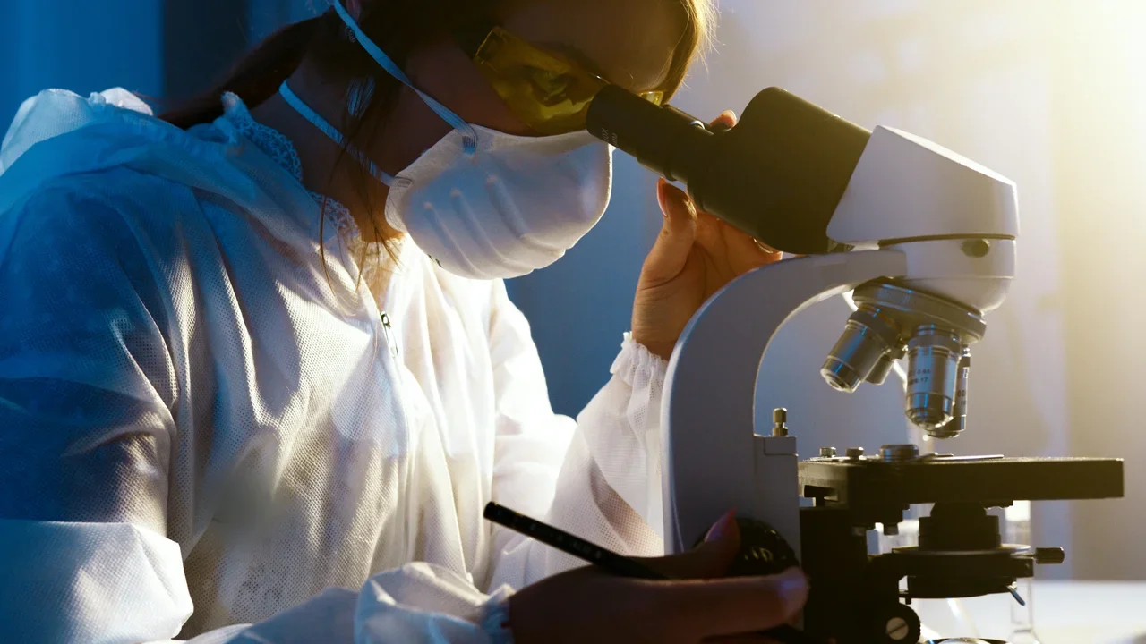 Scientist in protective gear looking into a microscope in a lab.